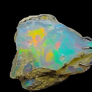 May include: A rough, natural opal gemstone with a milky white base and iridescent flashes of green, blue, yellow, and orange. The opal is held in a metal clamp.