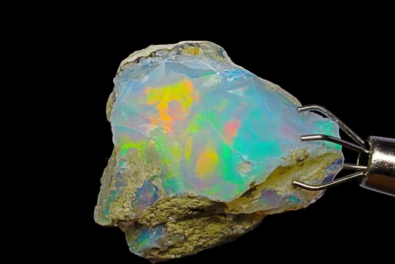 May include: A rough, natural opal gemstone with a milky white base and iridescent flashes of green, blue, yellow, and orange. The opal is held in a metal clamp.
