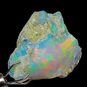 May include: A rough, natural opal gemstone with a blue, green, and orange iridescent sheen. The opal is held in a metal clamp.