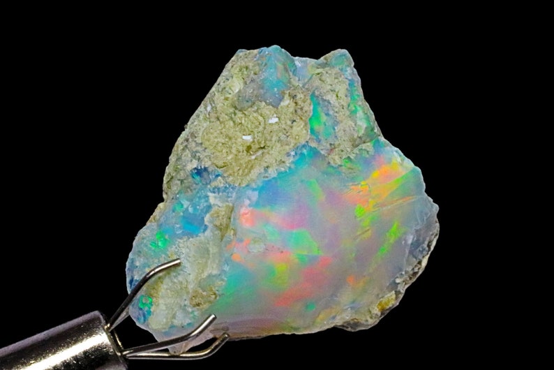 May include: A rough, natural opal gemstone with a blue, green, and orange iridescent sheen. The opal is held in a metal clamp.