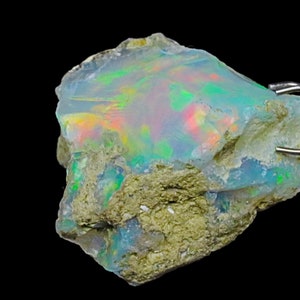 May include: A rough, natural opal gemstone with a milky white base and iridescent flashes of green, blue, and red. The opal is held in place by a metal clamp.