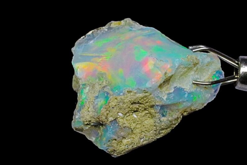 May include: A rough, natural opal gemstone with a milky white base and iridescent flashes of green, blue, and red. The opal is held in place by a metal clamp.
