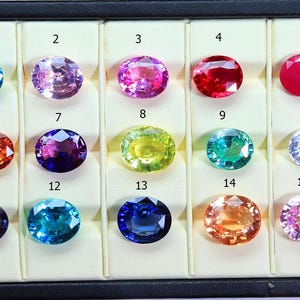 Peut inclure: Présentation de quinze pierres précieuses ovales dans un plateau noir avec des compartiments numérotés. Les gemmes varient en couleur, notamment bleu, rose, rouge, orange, vert et violet. Les pierres sont disposées en grille, mettant en valeur leurs teintes et leur clarté.