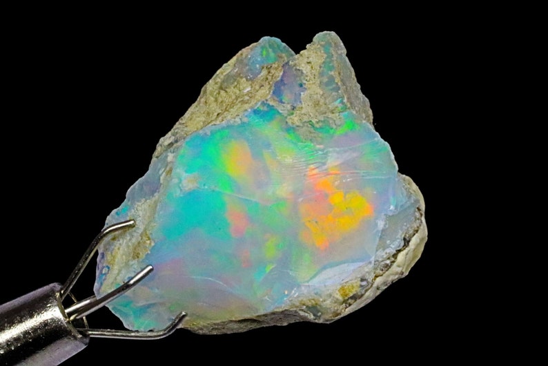 May include: A rough, natural opal gemstone with a milky white base and iridescent flashes of green, blue, orange, and yellow. The opal is held in a metal clamp.