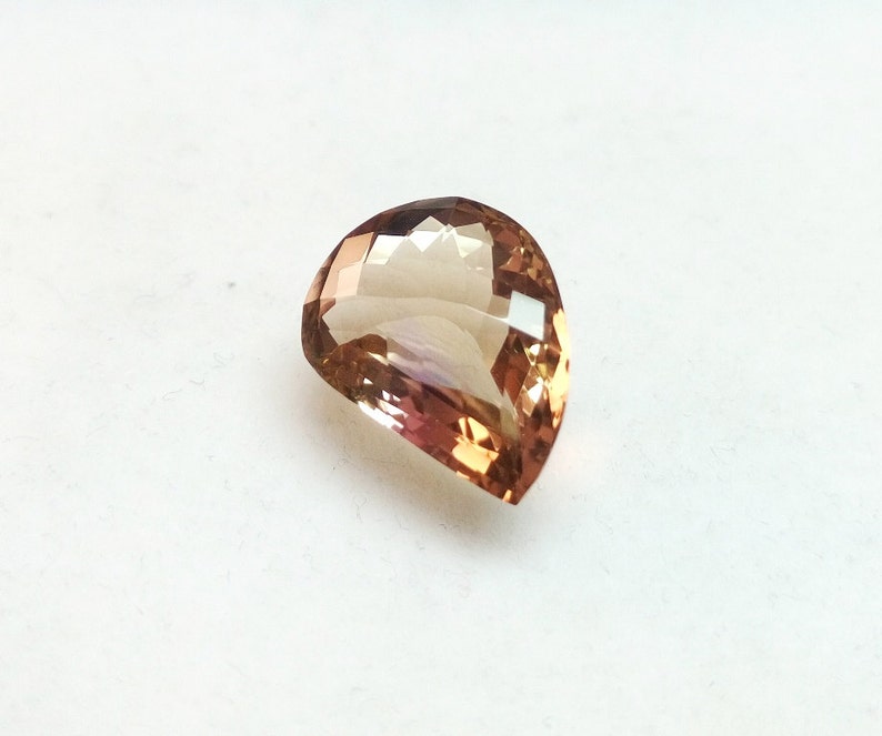 May include: A teardrop-shaped gemstone with a light brown to pink color gradient. The gemstone is faceted and has a high polish.
