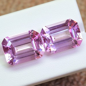 May include: Two pink emerald cut gemstones on a white display surface.
