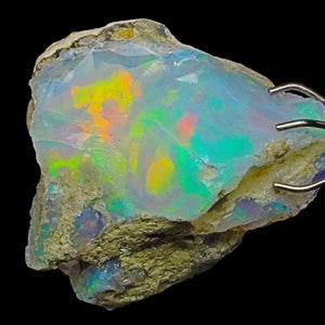 May include: A rough, natural opal with a milky white base and iridescent flashes of green, blue, yellow, and orange. The opal is held in a metal clamp.