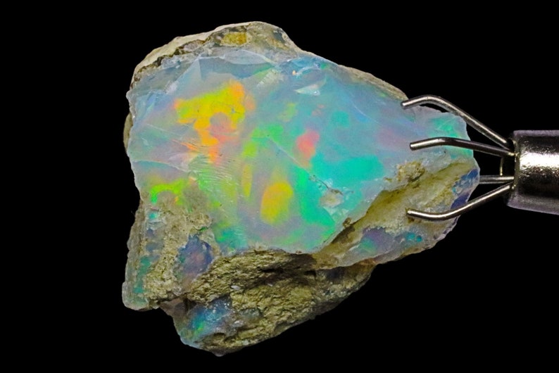 May include: A rough, natural opal with a milky white base and iridescent flashes of green, blue, yellow, and orange. The opal is held in a metal clamp.