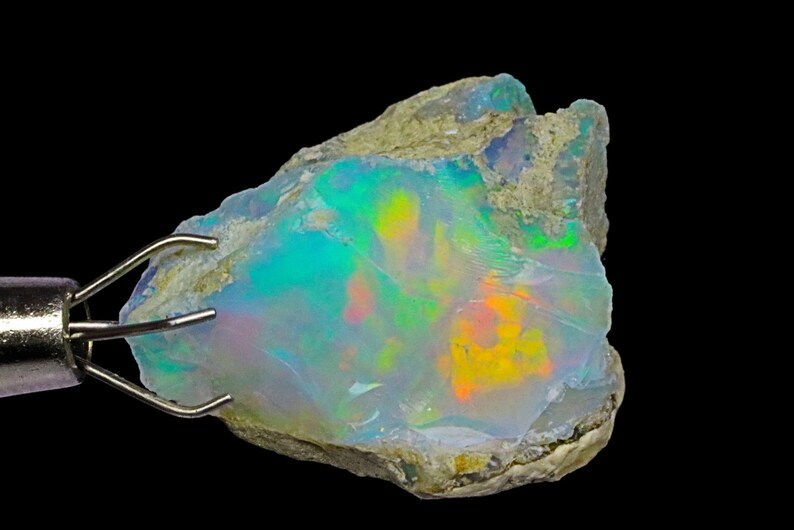 May include: A rough, natural opal gemstone with a milky white base and iridescent flashes of green, blue, yellow, and orange. The opal is held in a metal clamp.