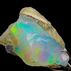 May include: A rough, natural opal gemstone with a milky white base and iridescent flashes of green, blue, and pink. The opal is set in a matrix of gray and brown rock.