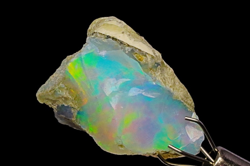 May include: A rough, natural opal gemstone with a milky white base and iridescent flashes of green, blue, and pink. The opal is set in a matrix of gray and brown rock.