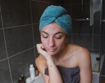 Può includere: Una persona con un asciugamano blu avvolto intorno alla testa si trova in un bagno. Sta guardando in basso e ha la mano sul mento. La persona indossa una parte superiore con motivo bianco e nero.
