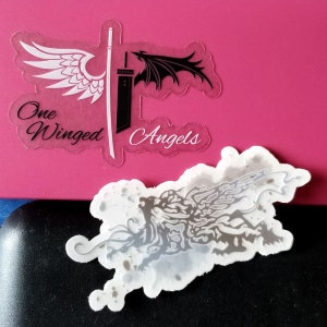May include: Two clear stickers with fantasy art. One features a white wing and a black wing with a sword, and the text "One Winged Angels". The other sticker has a gray illustration of a creature.