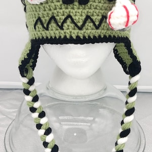 May include: A handmade crochet hat in a zombie theme. The hat features a green body with black accents, a black top, and a twisted cord with tassels. It has a white eyeball and a red and white eyeball attached.