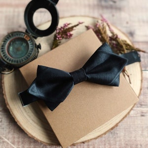 May include: A black bow tie presented on a brown paper box with a wooden base. The box is surrounded by pink flowers.