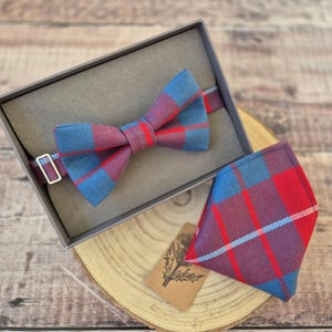 May include: A blue and red plaid bow tie and matching pocket square set. The bow tie is in a gray box. The set is displayed on a wooden surface. A small tag with a floral design is also present.