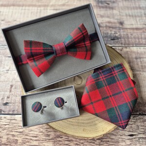 May include: A red, green, and navy plaid bow tie, necktie, and cufflinks set. The bow tie is in a gray box, and the necktie and cufflinks are on a wooden surface. The cufflinks are round with a plaid pattern.