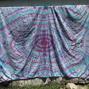 May include: A rectangular fabric tapestry featuring a tie-dye pattern with a central oval motif. The design displays a symmetrical arrangement of vibrant magenta, deep purple, and bright turquoise hues blended across the white cotton surface.