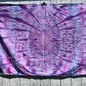 May include: A tie-dye textile in shades of purple, pink, and blue is displayed against a weathered wooden background. The fabric is draped, revealing a pattern of swirling colors. The textile is hung from a metal rod.