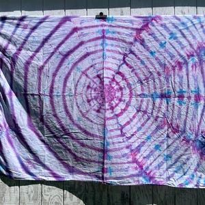 May include: A tie-dye textile in shades of purple, blue, and white, with a spiral pattern at the center. The fabric is hanging against a weathered wooden background, creating a vibrant contrast.
