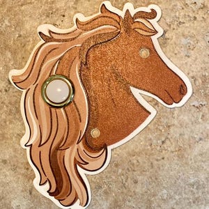 May include: A decorative horse head-shaped object with a light brown mane and a copper-colored body. The horse head has a white and gold-colored button in the center. The object is on a light brown surface.