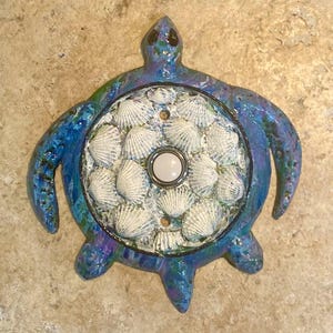 May include: A decorative light switch cover in the shape of a sea turtle. The turtle is painted blue with a shell-covered center and a white button. The switch plate is designed to look like a turtle.