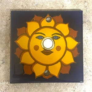 May include: A ceramic doorbell button with a sun face design. The sun is yellow with a brown outline and has a white button in the center. The background is dark blue.