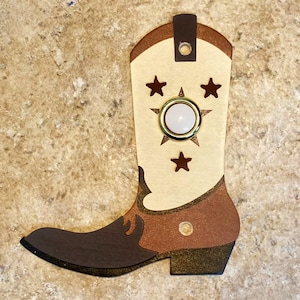 May include: A decorative cowboy boot cutout in shades of brown, tan, and cream. The boot features star accents and a central circular embellishment. A small brown loop is at the top.
