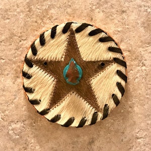 May include: A round, brown leather coaster with a black and white striped border. A five-pointed star made of brown leather is in the center of the coaster. The star has a turquoise stone in the center.