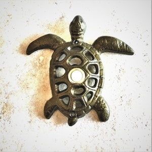 May include: A cast iron door knocker shaped like a turtle with a round, white button in the center. The turtle is painted in a dark bronze color.