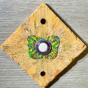 May include: A diamond-shaped doorbell button featuring a textured, light tan and gold painted finish with two mounting holes. Centered on the plate is a butterfly-shaped accent piece with a vibrant, multi-colored splatter pattern and a white button.