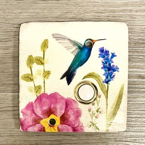 May include: A square stone light switch cover featuring a printed design of a blue hummingbird in flight, a large pink flower with a yellow center, and a tall stalk of blue flowers. A circular white toggle switch with a gold rim is centered.