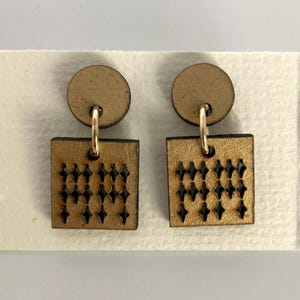 May include: A pair of gold-colored earrings. Each earring features a small, round top connected to a square pendant with a cut-out diamond pattern. The earrings are displayed on a white card.
