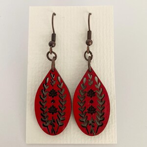 May include: A pair of red teardrop earrings with a floral design. The earrings have a dark floral pattern cut out of the red wood. The earrings hang from copper-colored hooks.