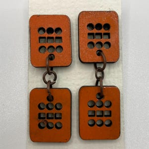 May include: Orange, square-shaped earrings with a geometric design of circles and squares. The earrings are connected by small, copper-colored rings. The earrings are displayed on a white card.