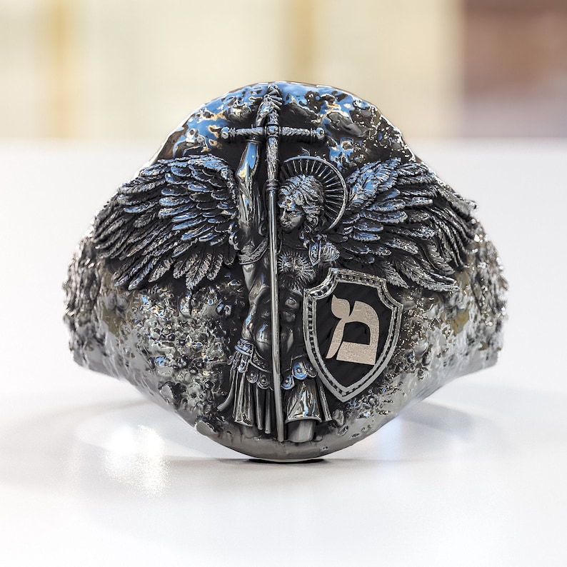 May include: A silver ring with a detailed design of an angel holding a sword and a shield with the Hebrew letter 'Dalet' on it.