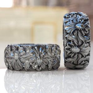 May include: Two silver rings with intricate floral designs. The rings are made of a smooth, shiny metal and have a detailed, three-dimensional pattern.