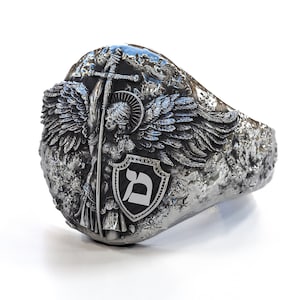 May include: A silver ring with a detailed design of an angel with wings holding a sword and a shield with the Hebrew letter 'Dalet' on it.