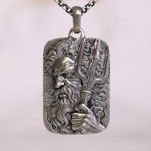 May include: A silver pendant necklace featuring a rectangular pendant with a detailed relief of a bearded man holding a trident. The pendant is attached to a silver chain. The design is intricate and the metal has a slightly aged appearance.