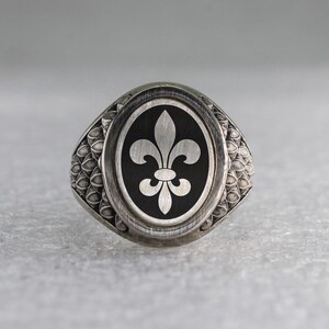 Könnte beinhalten: Ein silberner Ring mit einer schwarzen ovalen Einlage, die ein weißes Fleur-de-lis-Design zeigt. Der Ringband hat ein strukturiertes Muster.