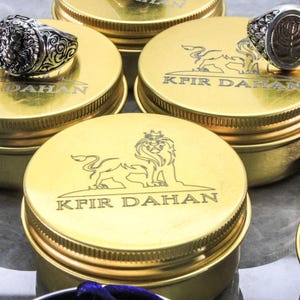 May include: Close-up of several gold-colored containers with screw-on lids. Each lid features a lion emblem and the text "KFIR DAHAN". Two silver rings with detailed designs rest on top of the containers. The rings have intricate patterns.