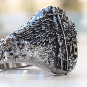 May include: A silver ring with a detailed design of an angel with wings, holding a sword and standing on a shield with a letter 'B' on it.