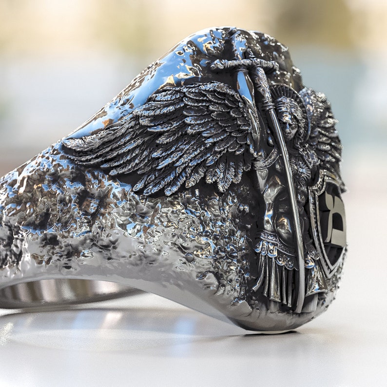 May include: A silver ring with a detailed design of an angel with wings, holding a sword and standing on a shield with a letter 'B' on it.