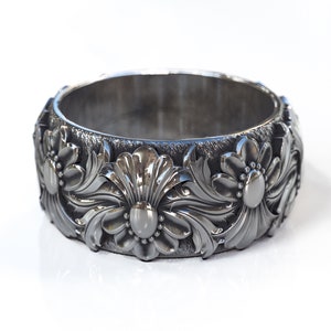 May include: A black metal ring with a floral design. The ring is wide and has a textured surface.