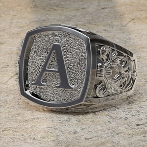 Könnte beinhalten: Ein silberner Siegelring mit einem erhabenen Buchstaben 'A' in der Mitte. Der Ring hat ein detailliertes, verziertes Design an der Band.