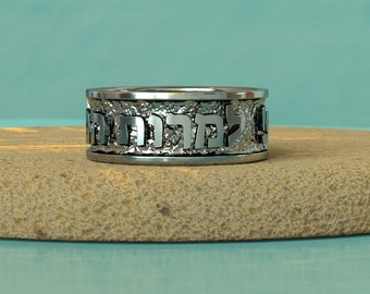 May include: A silver ring with Hebrew lettering inscribed around the band. The ring is sitting on a light brown textured surface.