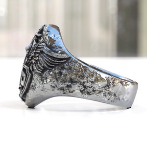 May include: A silver ring with a textured finish and a detailed design featuring a winged creature and a shield.