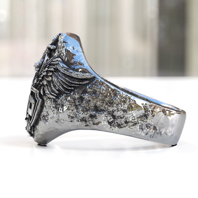 May include: A silver ring with a textured finish and a detailed design featuring a winged creature and a shield.