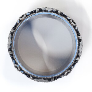 May include: A silver ring with a textured, ornate design around the band. The ring is circular and has a smooth, reflective surface.