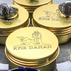 May include: Gold-colored metal boxes with a lion logo and the text "Kfir Dahan" engraved on the lid. The boxes are arranged on a white surface. A silver ring with a lion head design is on top of one of the boxes.